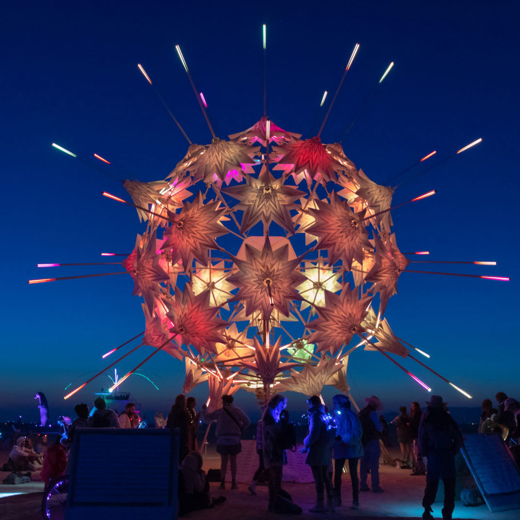 An art installation from Burning Man. It is a floating metallic ball, composed of lots of star shaped segments coloured orange, red and purple. There are black poles coming from the pole, whose tips are coloured purple, aqua green and orange. There is a crowd of people looking up at it as it floats in the night sky. 