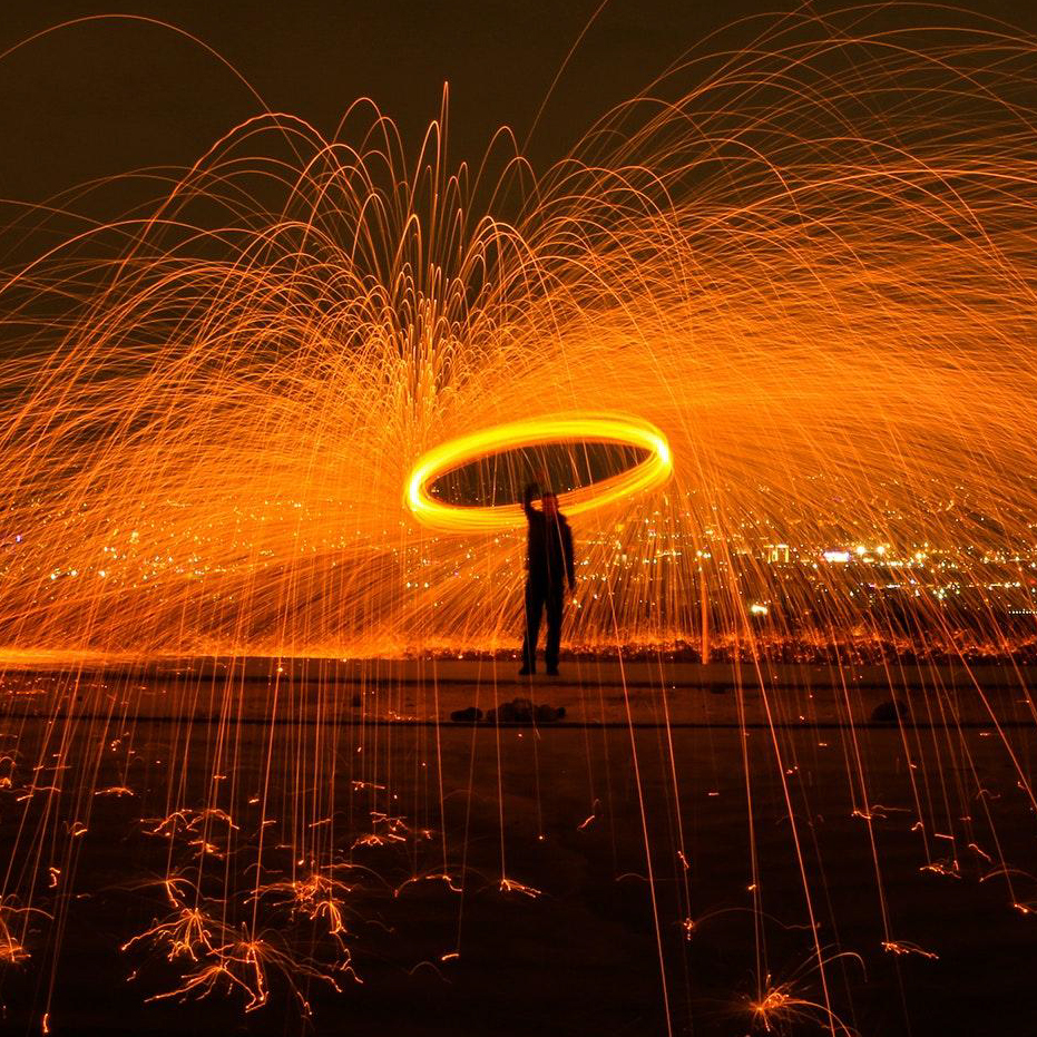 An artists spinning around a giant sparkler in the darkness, with lots of red strobes of light coming from it.The sparker is giving off a ring effect as he moves it around above his head. The ring is golden in colour and looks very hot.  